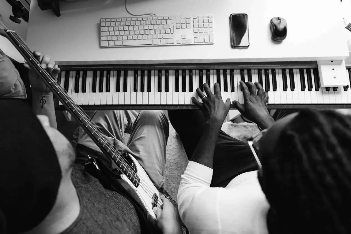 Two artists jam together, one plays a digital piano while the other a four-string electric bass. Both are sitting on the piano bench and the shot is taken from above.
