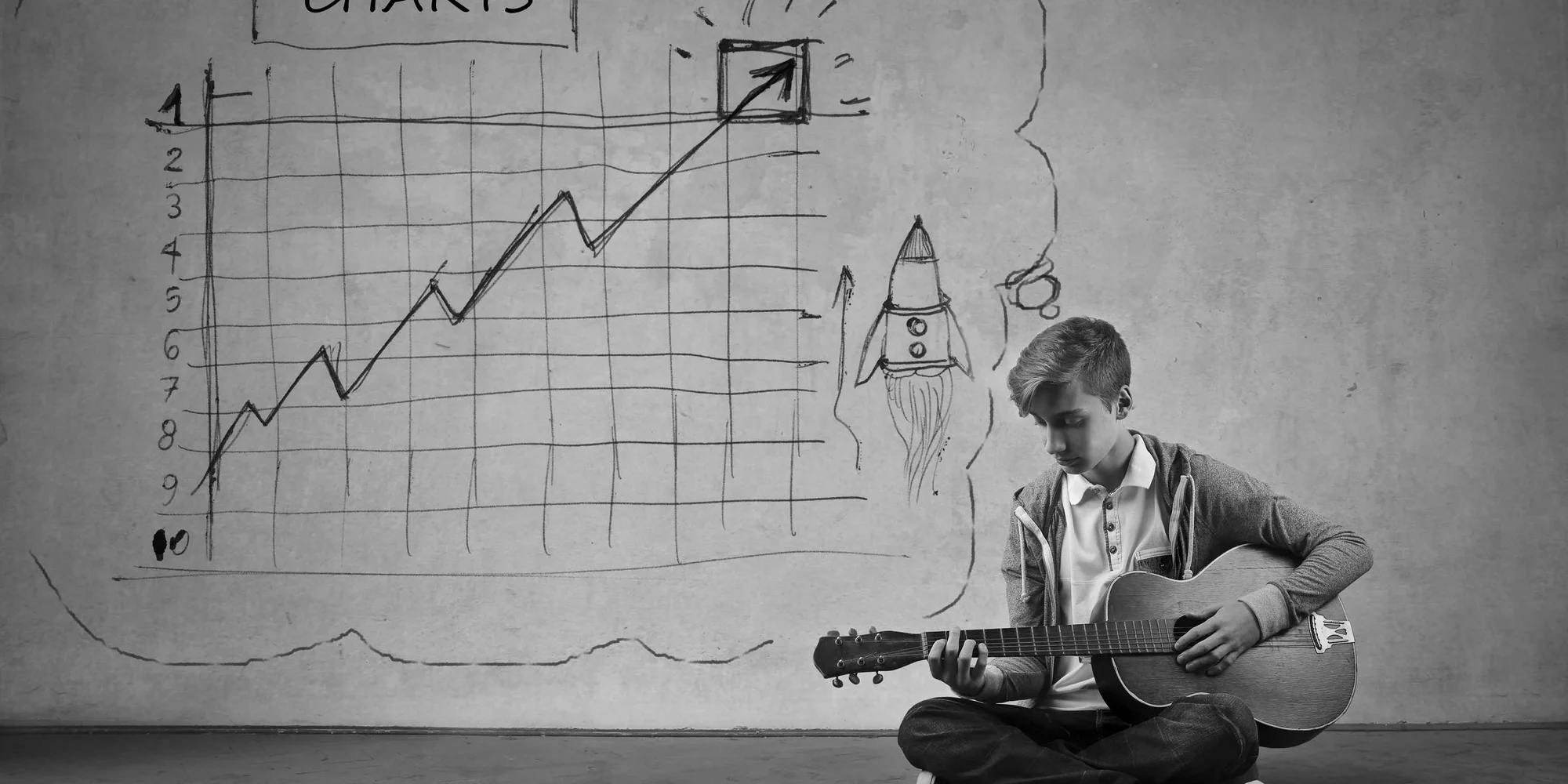 A male teenager is sitting on the floor playing an acoustic guitar in front of a chart that points up and has a rocket launching to the right of it.